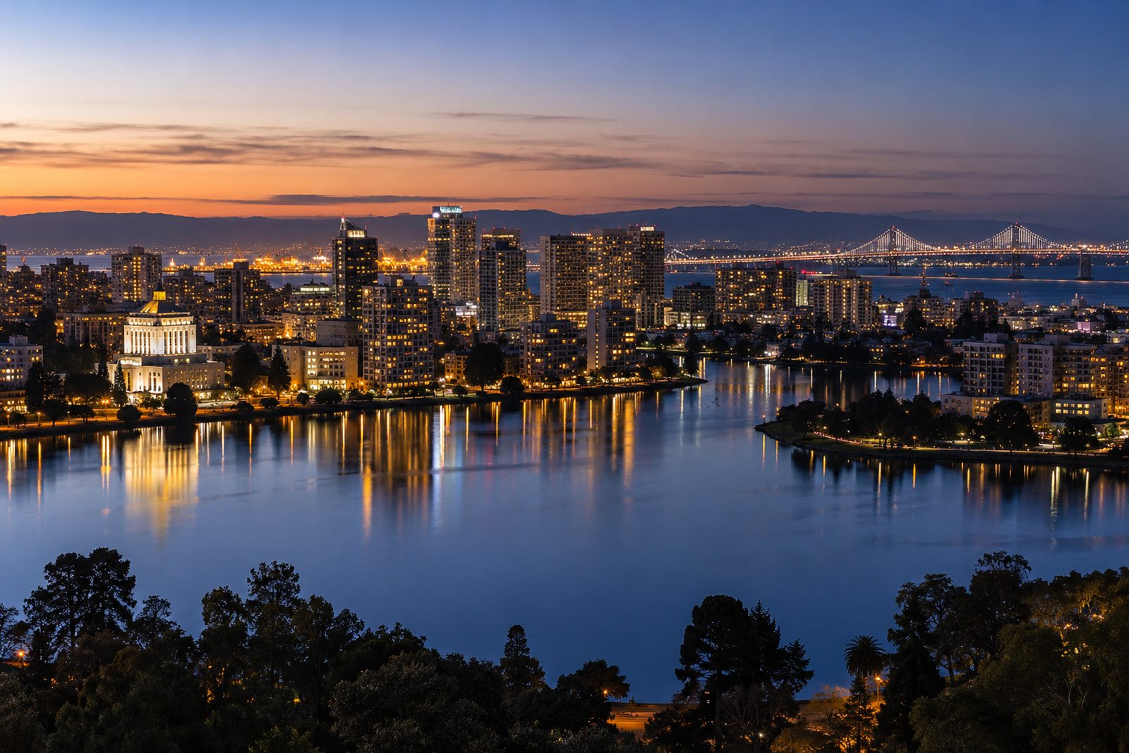 Oakland skyline over Lake Merritt at dusk with the Bay Bridge lit in the distance