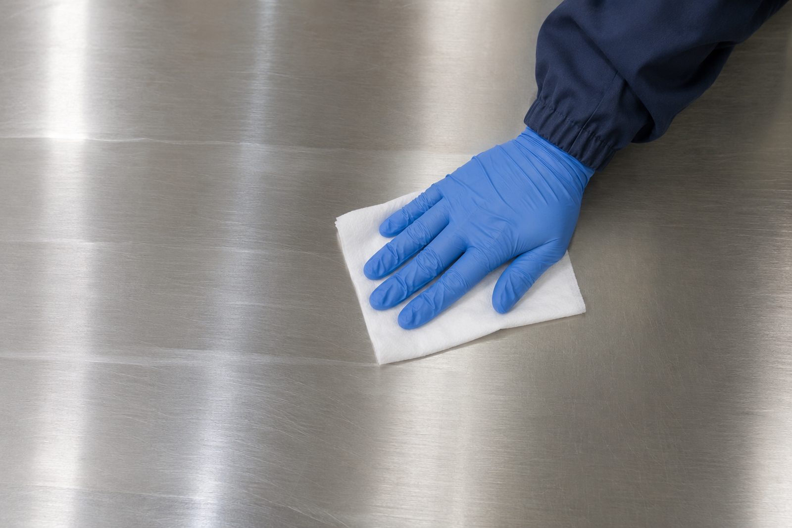 Top-down view of a gloved hand wiping a stainless steel lab bench with parallel overlapping strokes